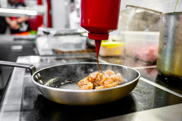 Close-up of a chef’s hand seasoning cooked shrimp in a pan with sauce from a squeeze bottle, in a professional kitchen
