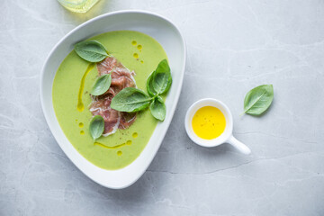 Bowl of green pea soup with torn prosciutto and basil, flat lay on a light-grey granite background, horizontal shot