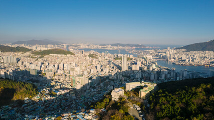an aerial view of Busan, Korea, showcasing dense urban areas with numerous buildings, surrounded by mountains and water bodies, under a clear sky.