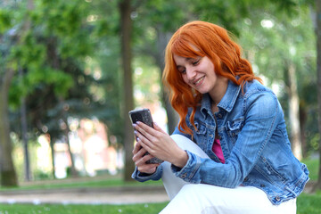 Young red haired woman smiling using mobile sitting in a natural space of the city, dressed casually, image with copy space. Lifestyle concept