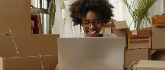A cheerful woman with glasses sits among cardboard boxes, happily working on her laptop, surrounded by a cozy indoor setting with plants.