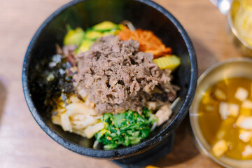 a traditional Korean dish, Bibimbap, served in a black bowl. It features colorful vegetables, beef, and a fried egg on top, garnished with sesame seeds.