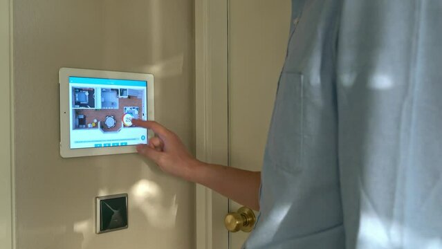 A smart home device is mounted on a wall next to a door. The device is displaying a video feed of a room. The room appears to be a living room with a couch and a chair
