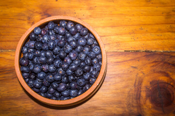 Lay flat of a rustic pottery bowl, filled with a large amount of Indigo c blueberries, on a wooden background