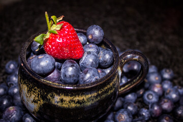 A single bright red Strawberry on top of a black tea-cup, filled with large indigo colored Blue berries