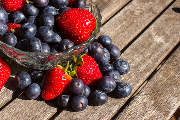 A glass bowl, filled to overflowing with bright red strawberries and indigo-colored blueberries on a weathered wooden surface