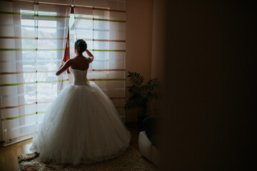 Fototapeta premium A bride stands by a window, veiled and in her wedding dress, gazing outside with a serene expression. The scene captures a moment of quiet reflection and anticipation before the ceremony.
