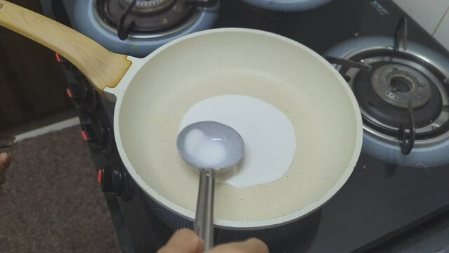 closeup a woman's hand pours white batter into a hot pan and spreading it using a ladle to make a round, delicious white dosa in the kitchen for breakfast under a gas stove