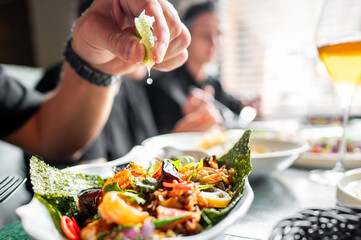 A hand holds a piece of food against a blurred background featuring vegetables and possibly seafood