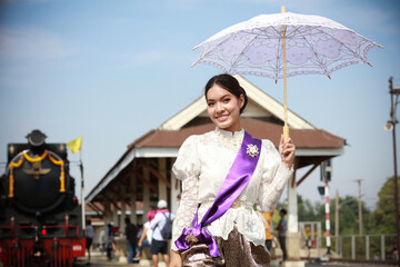 A portrait of a female in Thai traditional dress with umbrellas standing near a steam locomotive at the traditional train station.