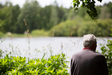 Back of older mans head and shoulders whilst  sitting down in countryside in front of a large lake with greenery and trees during summer