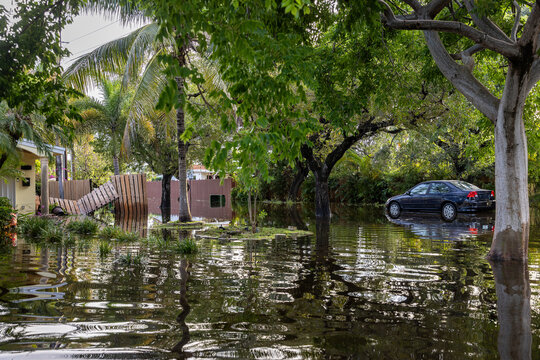 After the 2024 Hallandale Beach flood, a car is submerged in floodwaters outside a suburban home. Reflections in the water emphasize the severe impact on residential areas and the need for recovery.