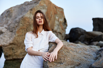 Serene woman in flowing white dress sitting on seaside rock with hands on hips gazing into the...