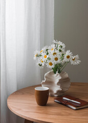 An aesthetic still life - a bouquet of daisies in a ceramic vase, a glass of coffee, a notebook in a leather cover on a round wooden table in the living room