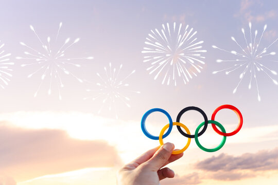 Hand holding Olympic rings against an evening sky, embellished with fireworks designs to evoke a celebratory atmosphere marking the start of the Olympic games