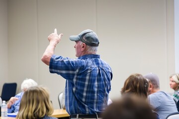 Senior man in a cap raising his hand to speak during a community meeting, engaging in a discussion with attendees in a casual setting.