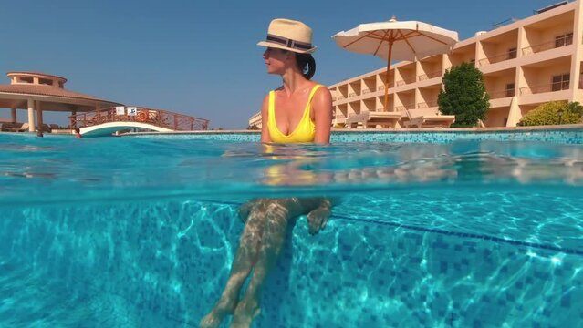 Girl in a hat sitting in the pool enjoys cool on a sunny day. Underwater split