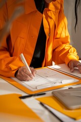 A woman in an orange trench coat sits at a table and writes in a notebook. Vertical orientation