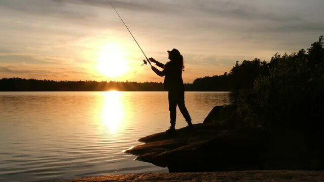 A woman cast a fishing line at sunset. The sun is setting in the background, casting a warm glow over the water. The woman is wearing a hat and is focused on her fishing rod. The scene is peaceful
