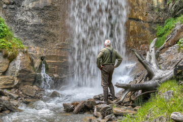Off the beaten path, an old, seasoned hiker stands in a ravine beneath a roaring waterfall. The thunder of the water fills the air as he marvels at the raw power of nature.