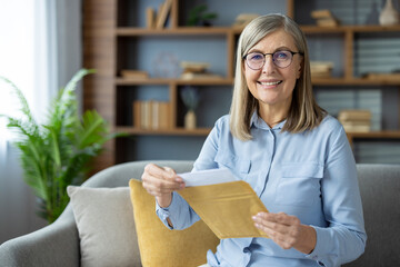 Happy senior woman sitting on the sofa at home, opening a yellow envelope with a smile and holding a letter.