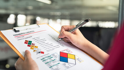 Action of a safety supervisor hand is checking on checklist of chemical and hazardous material form to verify the risk with blurred background of factory storage workplace. Industrial working scene.