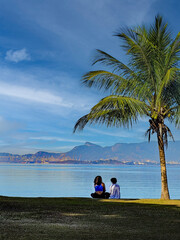 Two people sitting on the grass by a bay, chatting in a relaxed attitude. In the background is Corcovado Hill, where the statue of Christ the Redeemer stands.