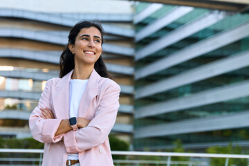 Confident portrait of young middle eastern Israel businesswoman standing with crossed hands at office building. Successful smiling indian or arabic woman in business suit looking aside. Copy space