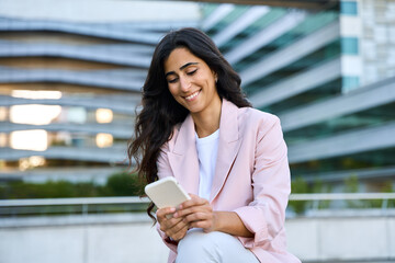 Smiling young middle eastern Israel businesswoman using smartphone mobile phone online app for work at office building. Successful 20s happy indian or arabic woman in business suit holding cellphone