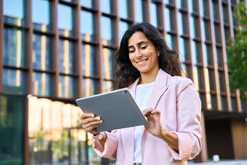 Smiling young middle eastern Israel businesswoman using tablet pc application for online remote work at office business building outdoors. Indian or arabic woman holding digital computer. Copy space