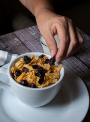 Concept of taking or grabbing food with hands, feminine Mexican hands. Putting nuts on cereal in a white cup on a white plate on an old wooden table, with a napkin next to it, vertical close up image