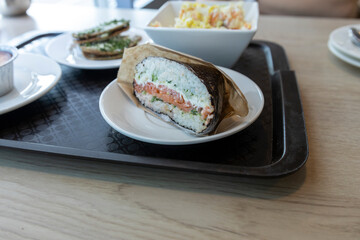 A close-up shot of a sushi hand roll filled with salmon and avocado, resting on a white plate. Its on a black tray with other dishes, showing a relaxed dining experience.