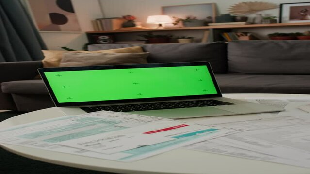 No people vertical shot of laptop with emty green chromakey screen, domestic bills, household invoices, tax return forms, on table in living room of family home. Suitable as template, copy space