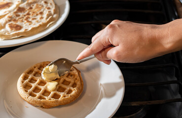 Concept of taking or grabbing food with hands, feminine Mexican hands spreading butter on a waffle with a spoon and on a white plate and a black table 
