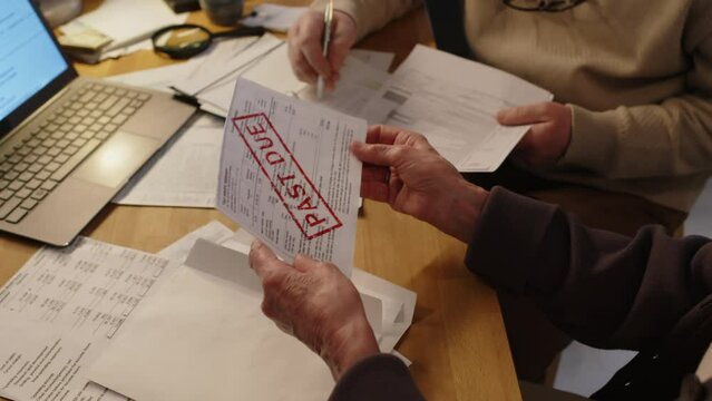 Close-up of hands of unrecognizable senior woman sitting at table with heap of invoices, taking unpaid bill with missed payment and red overdue stamp out of envelope and showing to husband