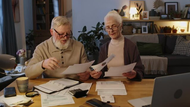 Medium arc shot of 70-year-old Caucasian husband and wife sitting in living room at home, examining bunch of utility invoices, medical expenses and mortgage bills, deciding what to pay first
