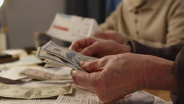 Close-up shot of wrinkled hands of anonymous elderly man studying electricity bill, and unrecognizable woman counting out American dollars, while managing family financial affairs