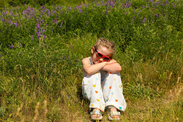 A little girl in sunglasses sits in a green meadow hugging her knees among grass and purple flowers.