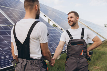 Two happy bearded workers in uniform talking and shaking hands near solar panel farm