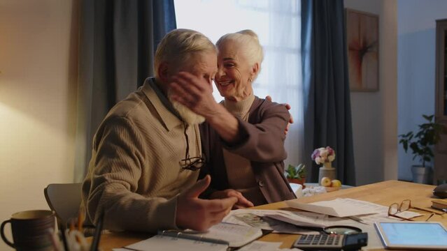 Medium shot of elderly Caucasian husband and wife sitting at table, sorting mail, woman opening envelope, taking out letter and cash or gift voucher prize, cheering and hugging man