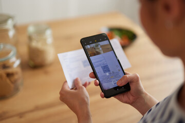 Asian woman standing in the kitchen with mobile phone paying bills online