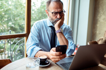 Concerned businessman checking phone while sitting at a cafe table with laptop and coffee cup