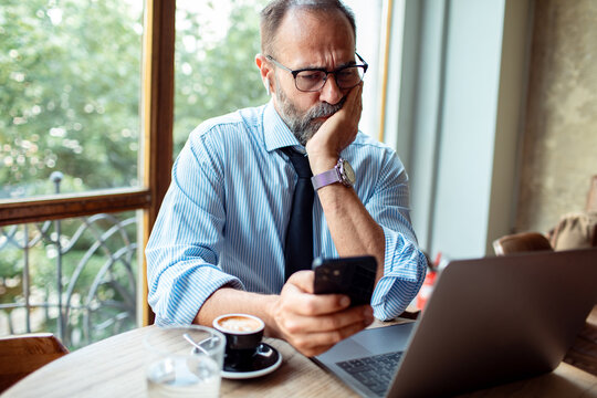 Concerned businessman checking phone while sitting at a cafe table with laptop and coffee cup