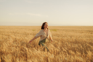 A happy young woman in casual attire enjoying a walk in a beautiful golden wheat field at sunset.