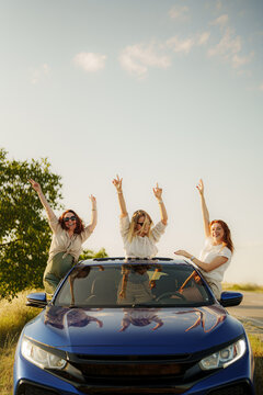 Three happy women with arms raised in excitement, standing through car sunroof on a sunny day, embodying freedom and joy during a road trip.