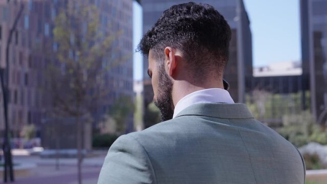Young handsome Caucasian business man walking talking on phone serious expression. Male professional in suit receiving mobile call stroll outdoor. Successful worker with background of buildings.