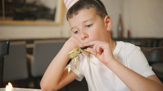 A boy in a white t-shirt looking sad and lonely,blowing a whistle, sitting alone at an empty table. He wears a party hat but seems disappointed