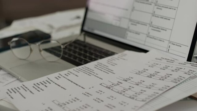 Close-up no people shot of bank savings account statement lying on top of laptop, with table from personal finance management program on screen, and eyeglasses on table at family home