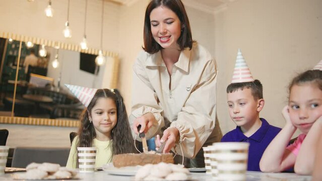 Smiling kind mother of the birthday boy cuts a birthday cake and puts pieces on the children's plates