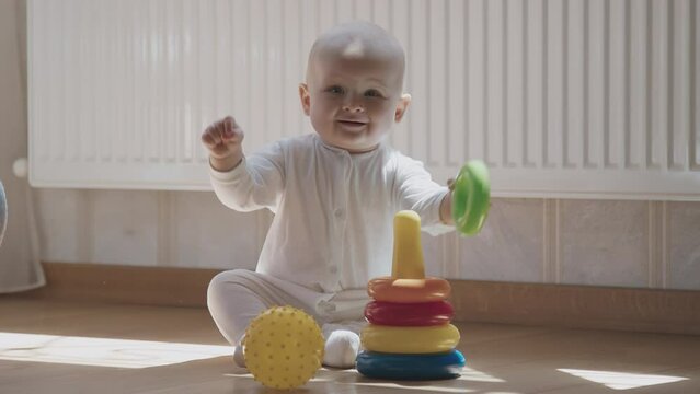 Fun Baby boy in white outfit sitting on wood floor, play with colorful stack rings and yellow spiky ball. Infant enjoy playtime with vibrant toys, showcase early child development and motor skills. 